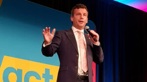 Act Party leader David Seymour, who proposed the Principles of the Treaty of Waitangi Bill, speaking on stage with a yellow Act sign behind him.